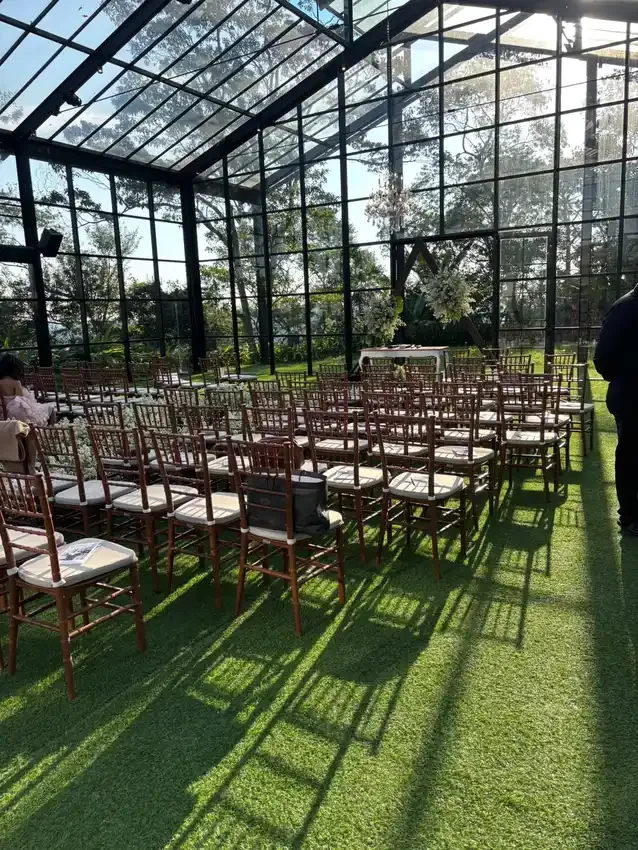 Wooden chairs lined up on a lawn inside a glass greenhouse, set up for a wedding ceremony in Brazil