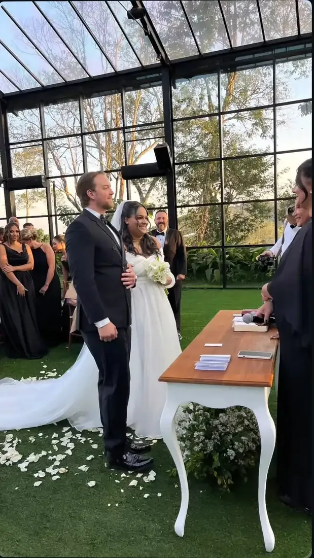 Couple at a bilingual wedding ceremony in a glass greenhouse, in front of the officiant's table, guests around