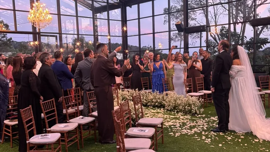 Cerimônia de casamento em salão envidraçado com convidados sentados e noivos no corredor decorado com flores