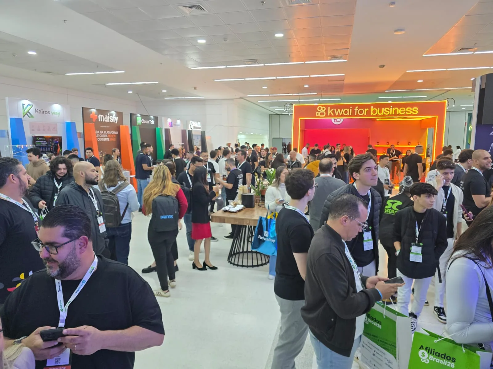 Visitantes com crachás circulando em evento de afiliados em São Paulo, com estandes de empresas ao fundo
