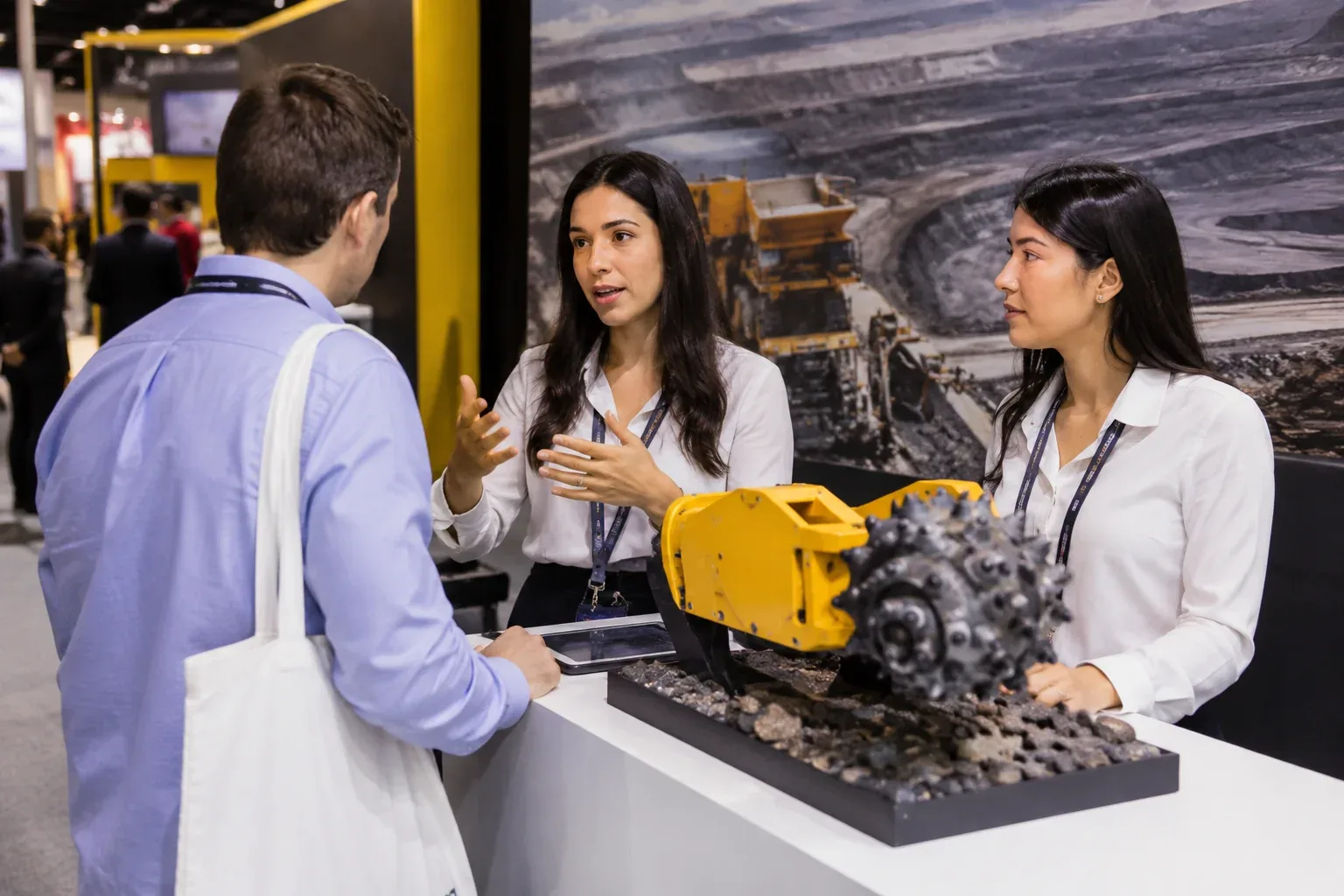 Intérprete conversando com visitante em estande de feira de mineração, ao lado de colega e de uma máquina em miniatura