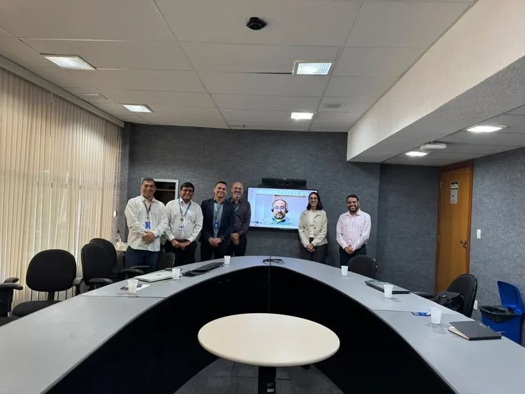 Reunião em Brasília com seis participantes em pé e um consultor indiano participando por videoconferência na tela